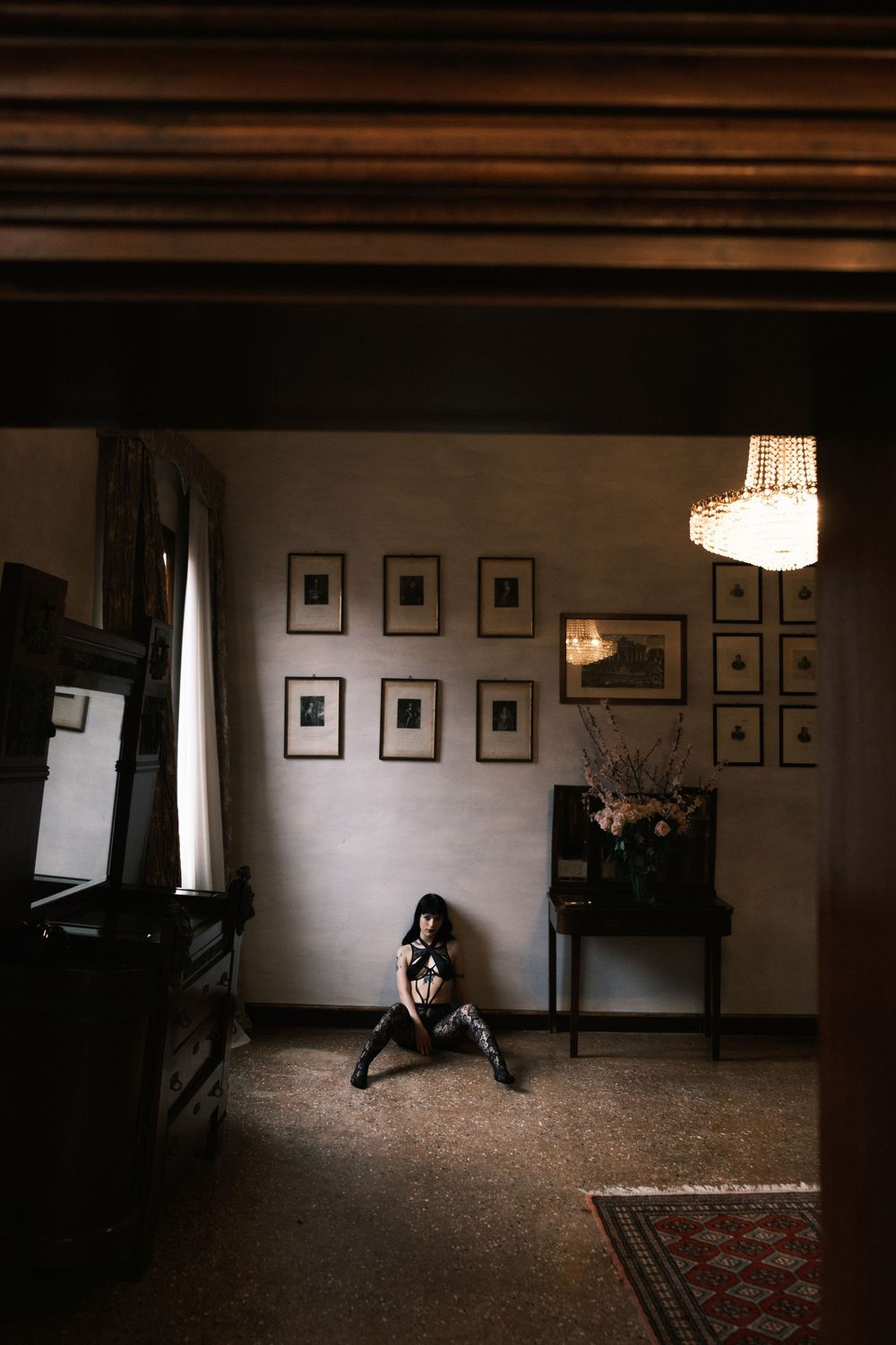A woman sits on the floor of a dimly lit vintage room with framed artwork on walls and a chandelier overhead.