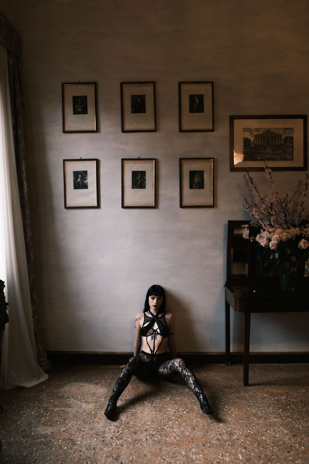Woman in black lingerie and boots sits against a gray wall with framed artwork in an elegant room.