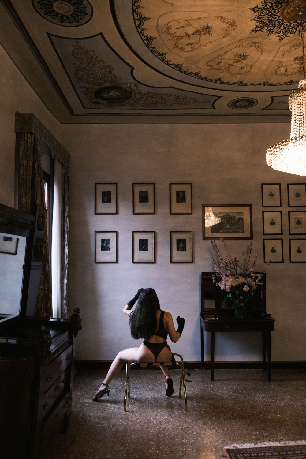 Woman in black lingerie sitting on chair in ornate room with framed artwork and decorative ceiling.