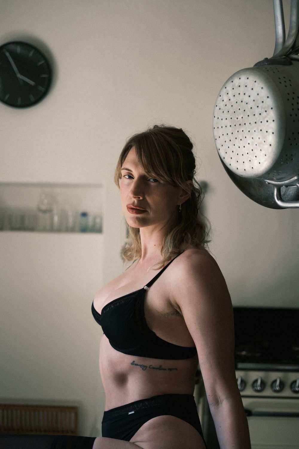 Woman in black lingerie standing in kitchen with clock and hanging colander in background.