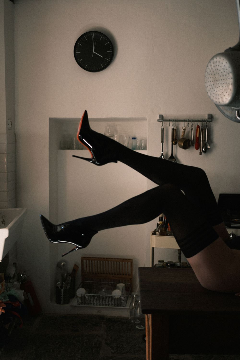 Person lying on kitchen counter with legs raised wearing black stockings and patent leather heels with red soles.