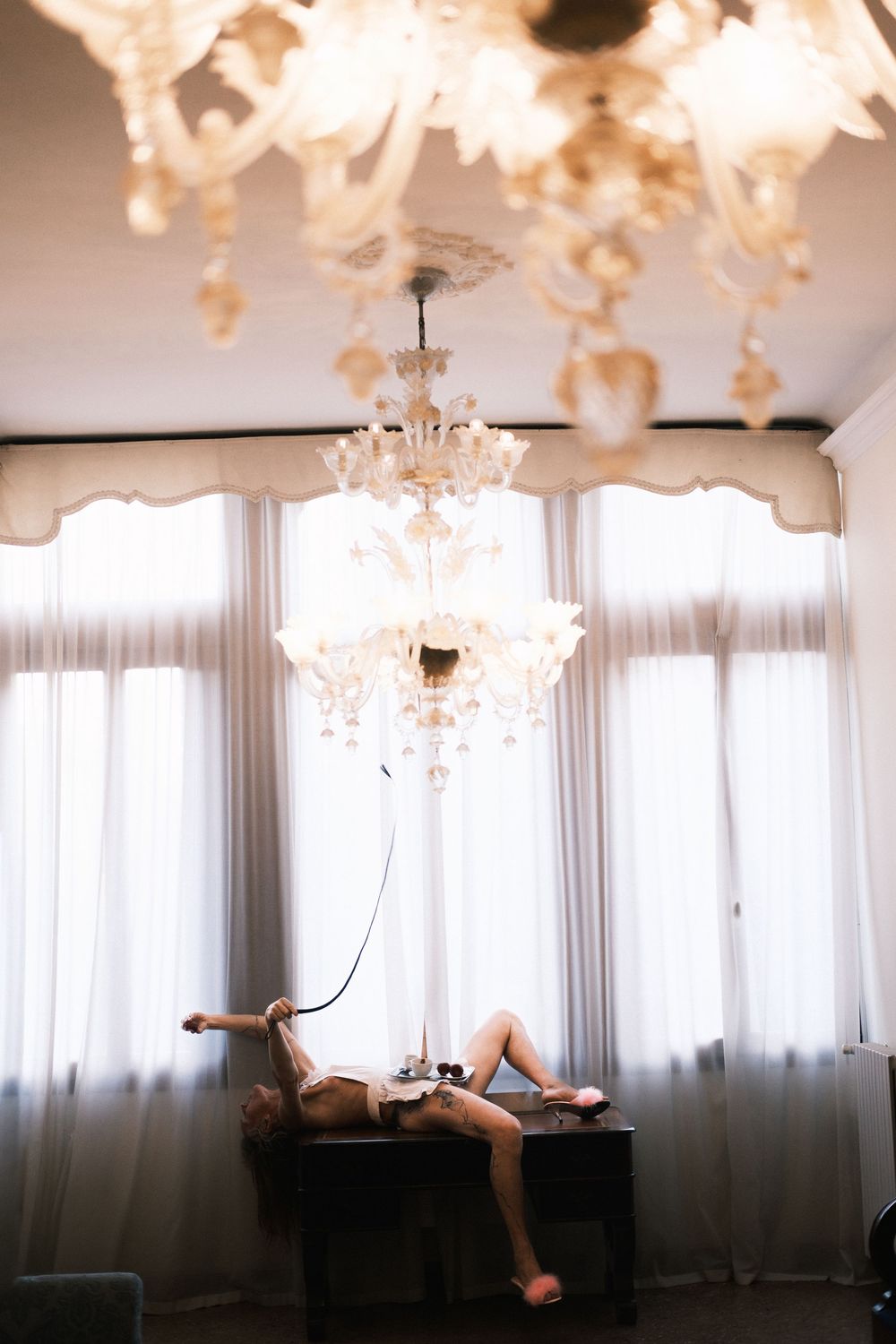 Elegant room with crystal chandelier, woman reclining on dark wooden table near bright curtained windows.
