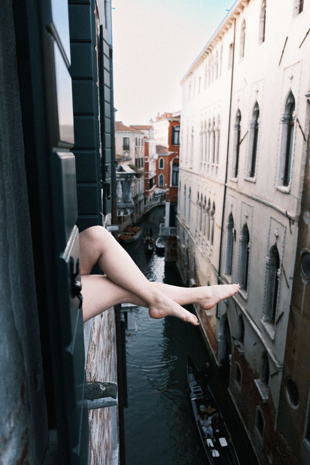 Bare legs dangling from a window overlooking a narrow Venice canal with historic buildings.