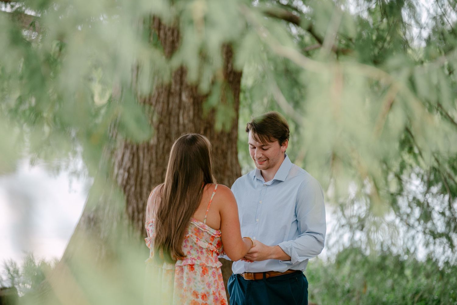 Couple sharing a romantic moment outdoors under a willow tree, man smiling at woman in floral dress.