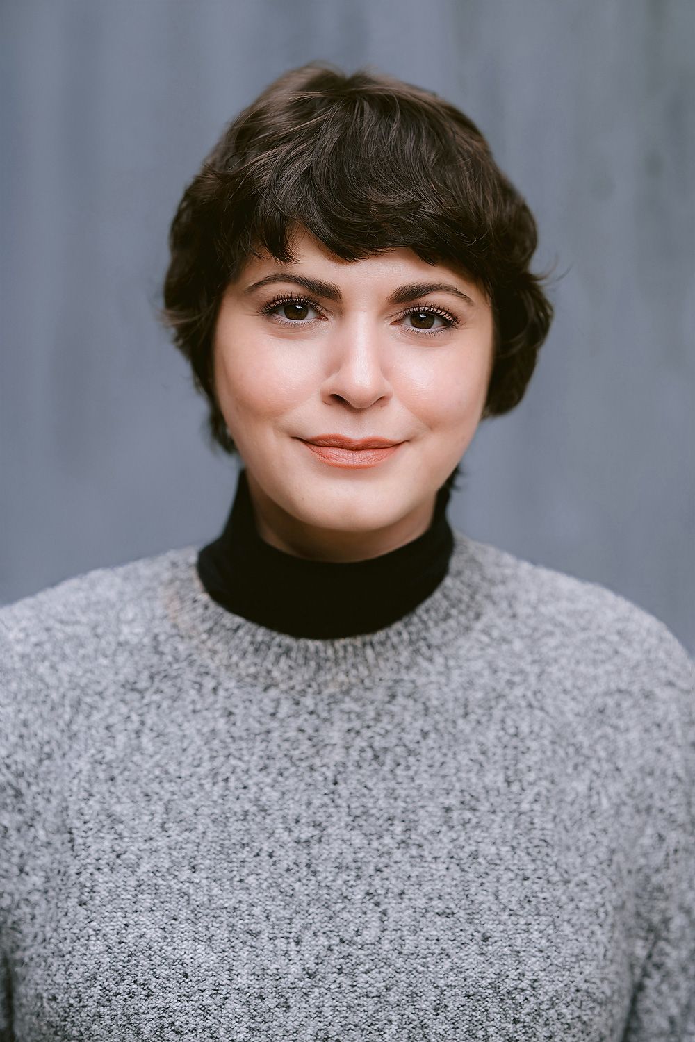 Young actress headshot with short dark hair wearing a gray turtleneck sweater, smiling softly against a gray background.