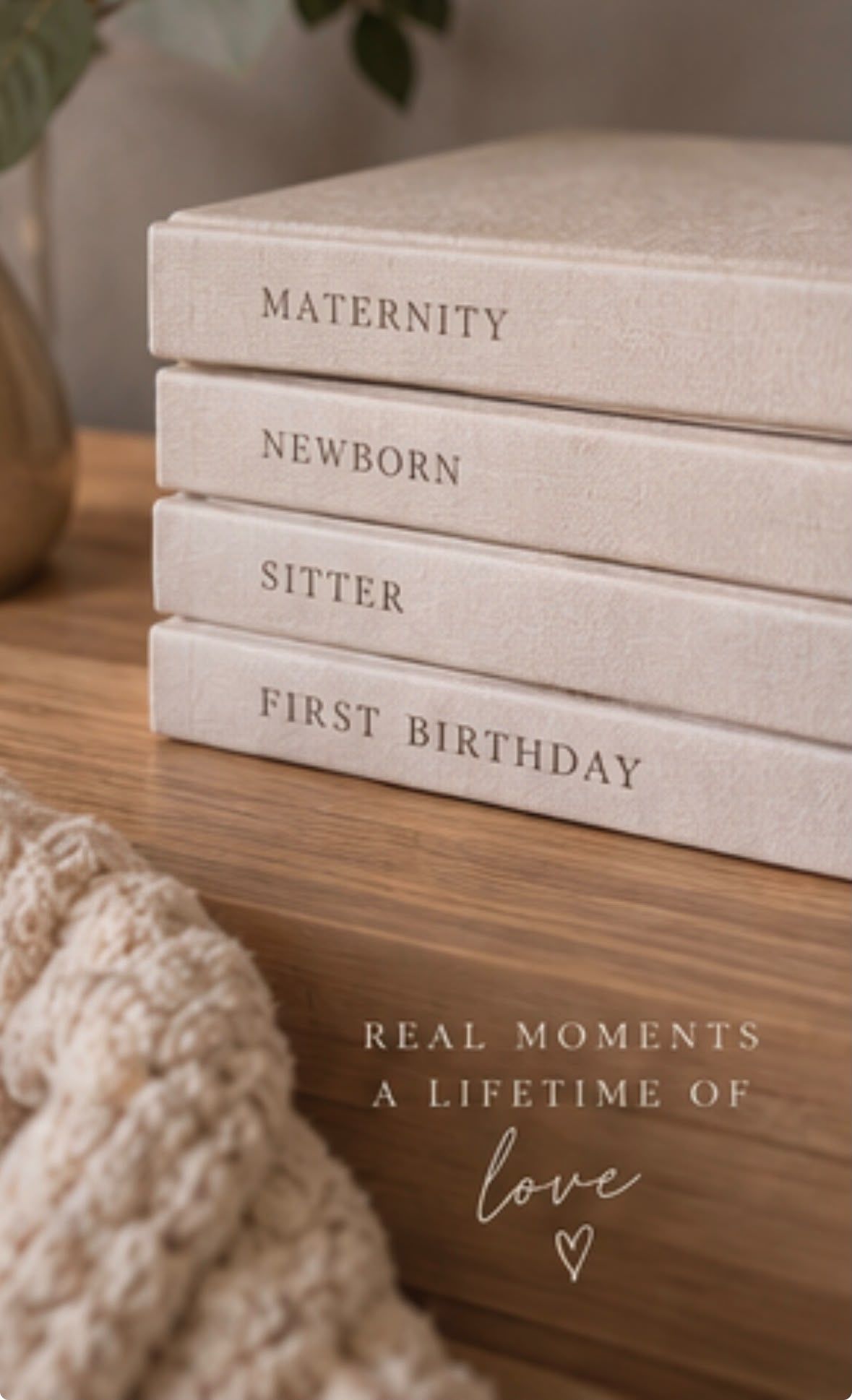Four stacked linen photo albums labeled Maternity, Newborn, Sitter, and First Birthday on a wooden shelf.