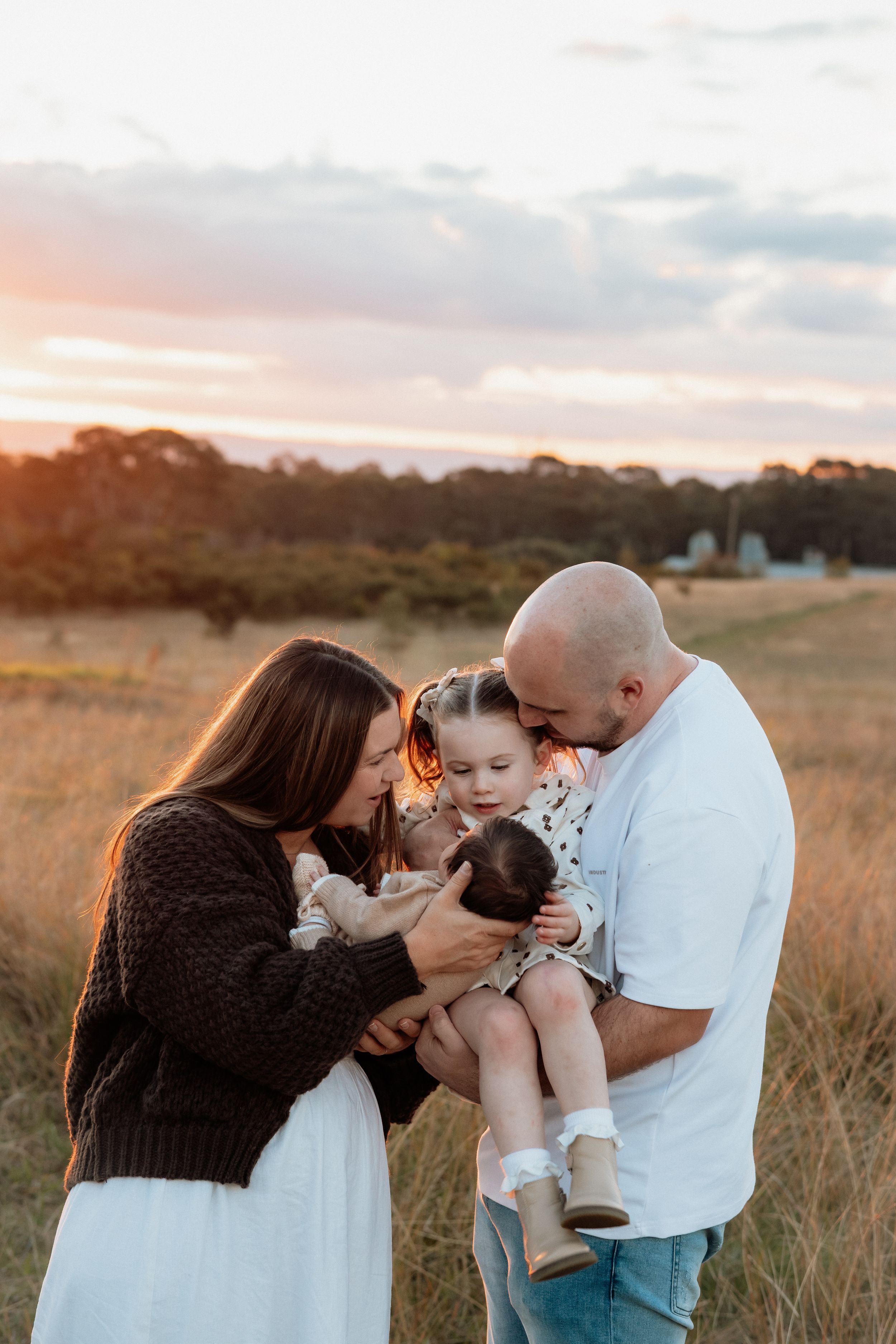 Family of four sharing a tender moment in a golden field at Rouse Hill Regional Park at sunset, parents kissing toddler and newborn baby.