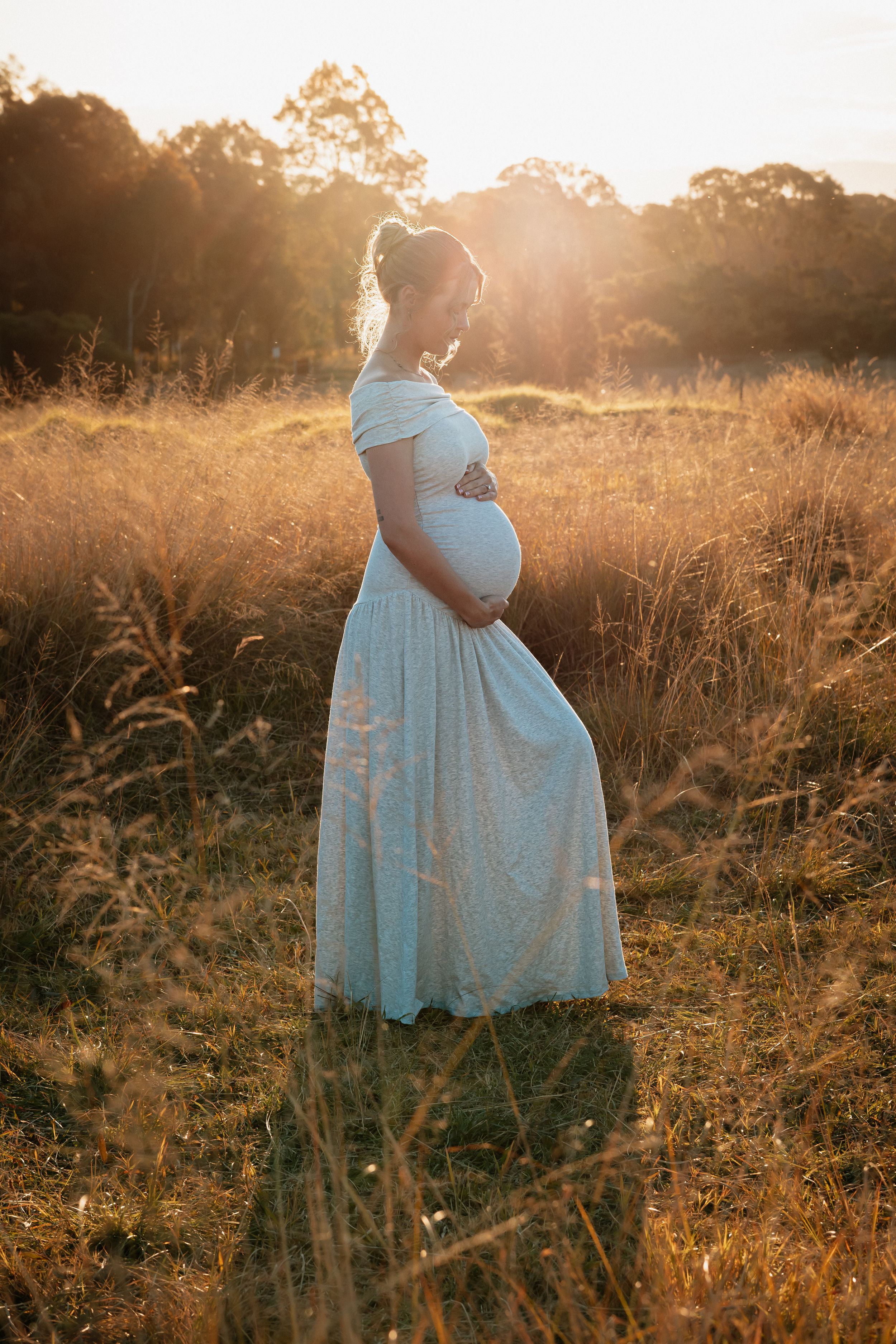 Pregnant woman in white lace dress cradling her baby bump in golden sunset field at Rouse Hill Regional Park during maternity photoshoot.