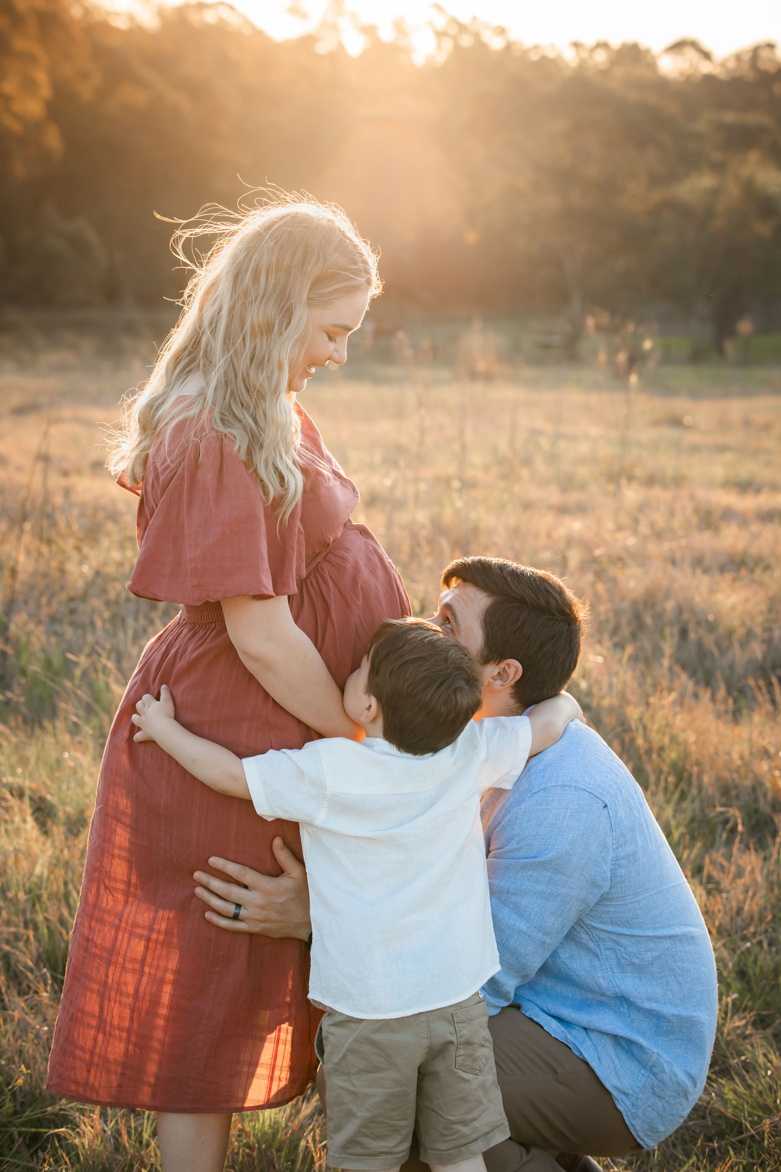 A pregnant woman in a red dress poses with her husband and young son in a golden sunset field in Rouse Hill for a maternity photo.
