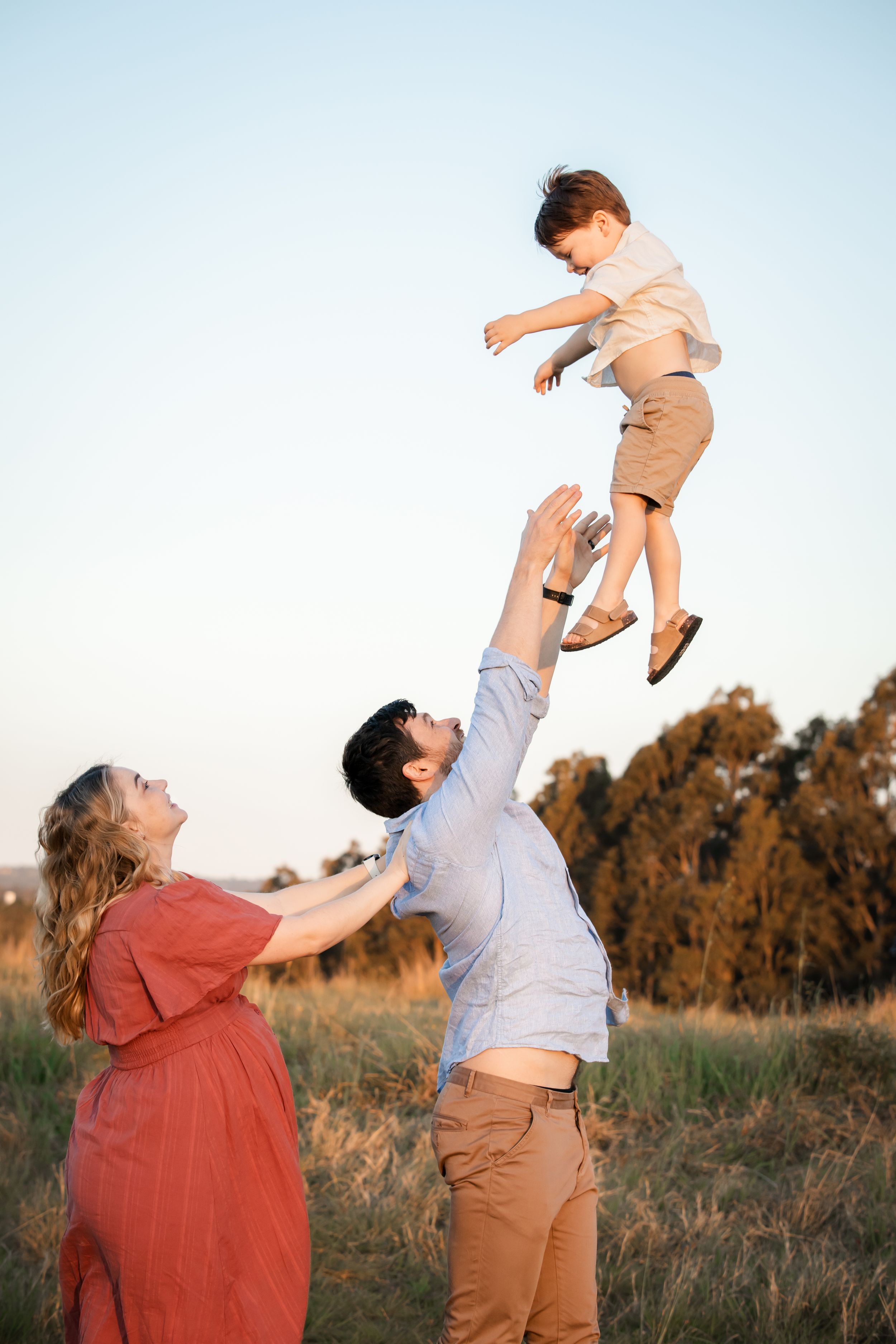 A joyful family outdoors in Rouse Hill as a father tosses his young son high in the air while a pregnant mother watches and smiles.