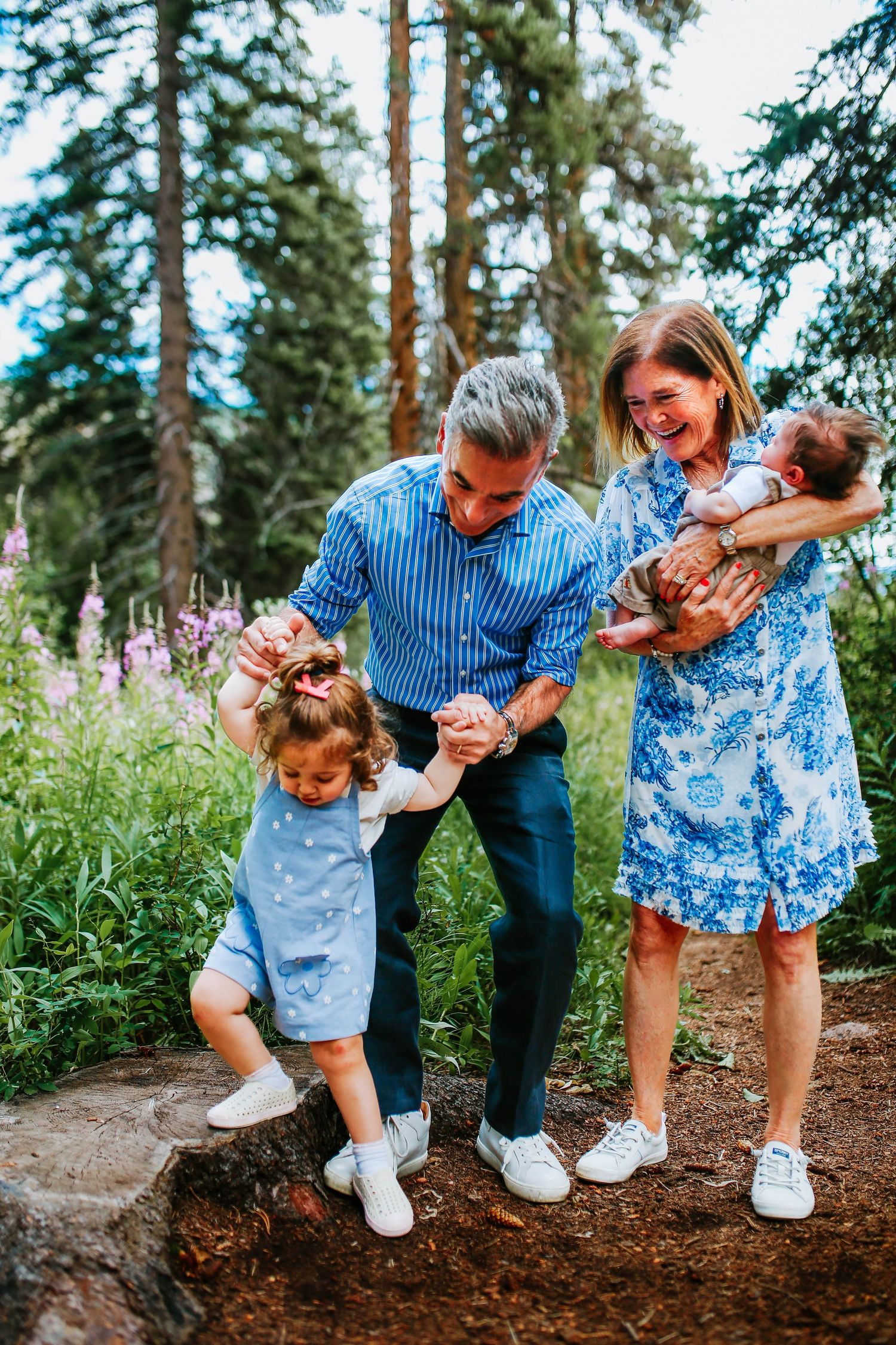 Family of four enjoying outdoor forest setting, parents playing with toddler on rocks while mother holds newborn baby.