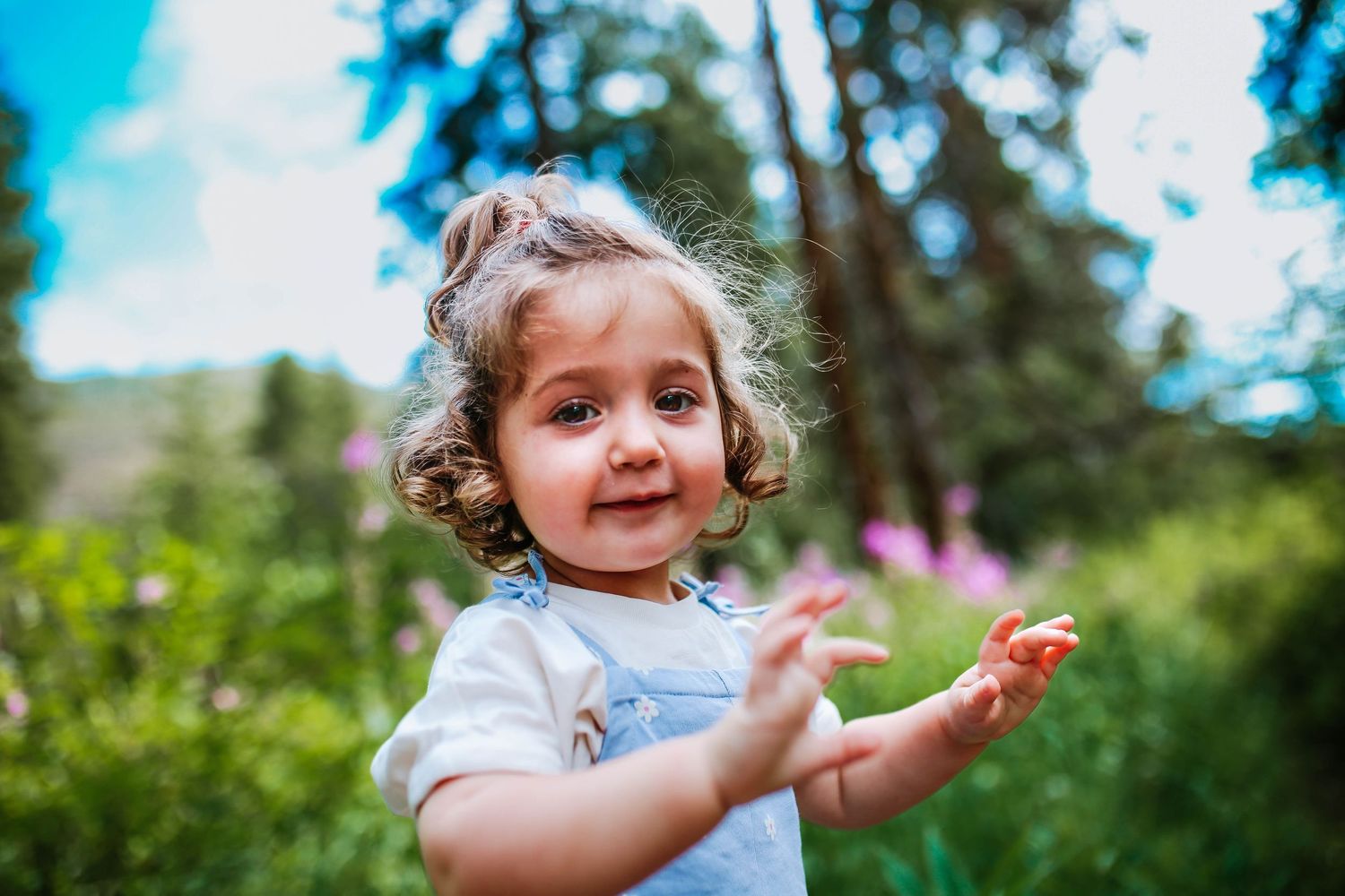 Adorable toddler girl with curly hair in denim overalls smiling outdoors in a lush green garden.