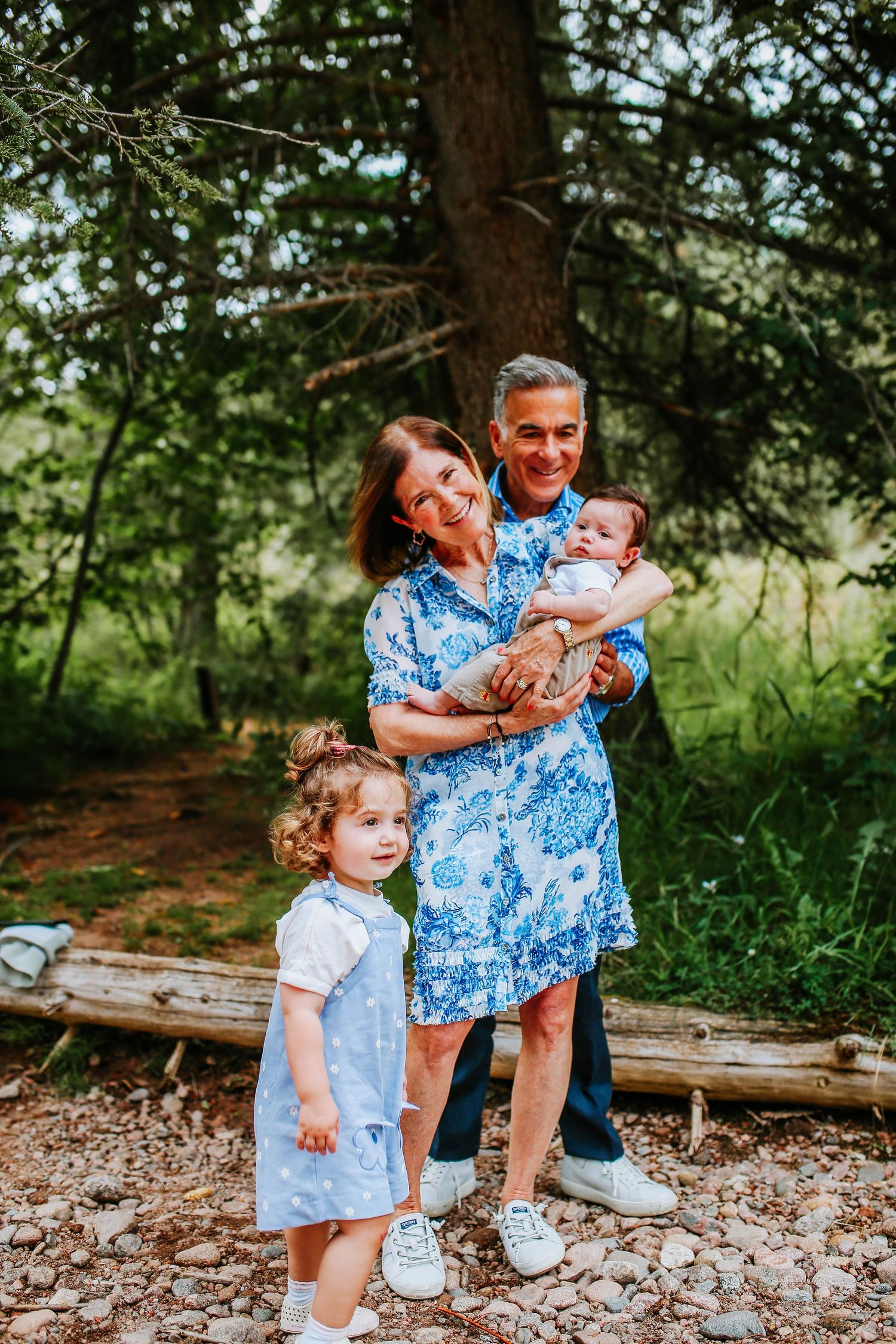 Happy grandparents posing outdoors with toddler girl and baby in arms, surrounded by lush green trees.