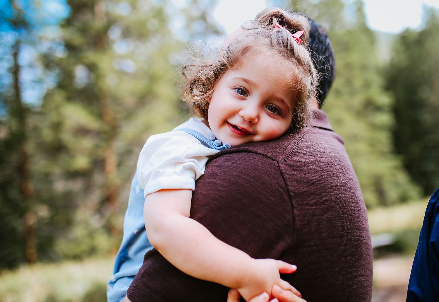 A smiling toddler girl with curly hair being held by an adult outdoors near green trees.