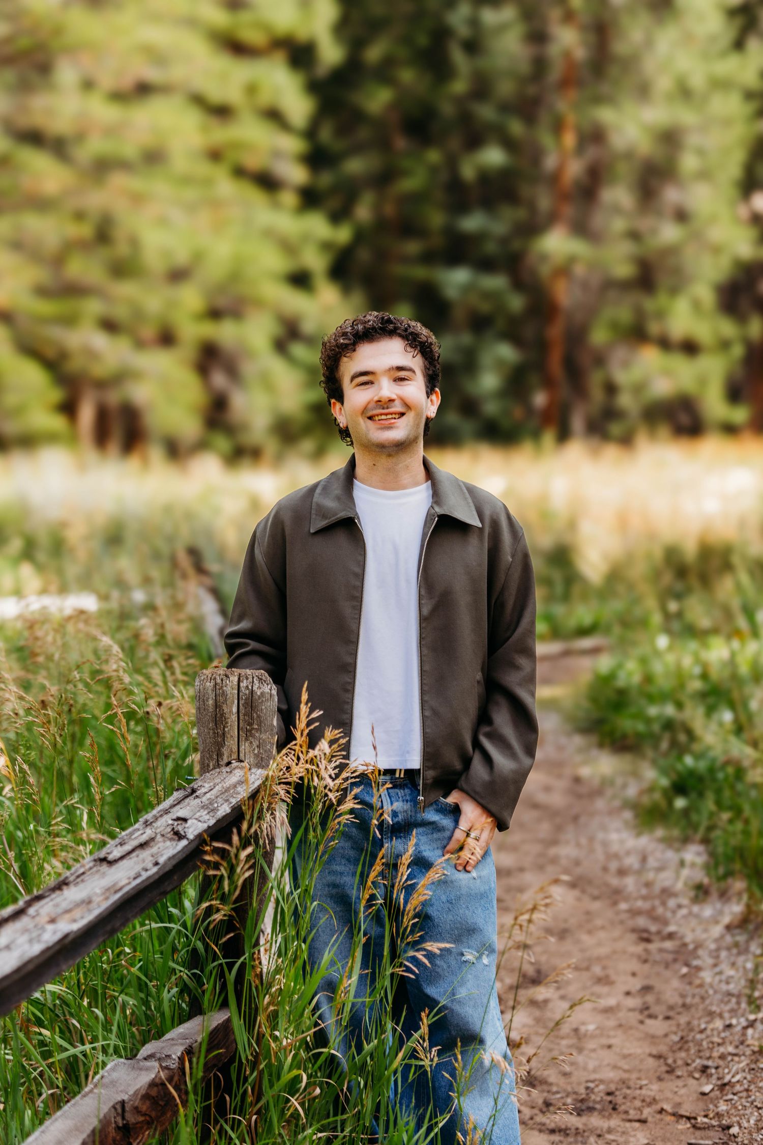Smiling young man in jeans and dark jacket leaning on a wooden fence along a forest trail.
