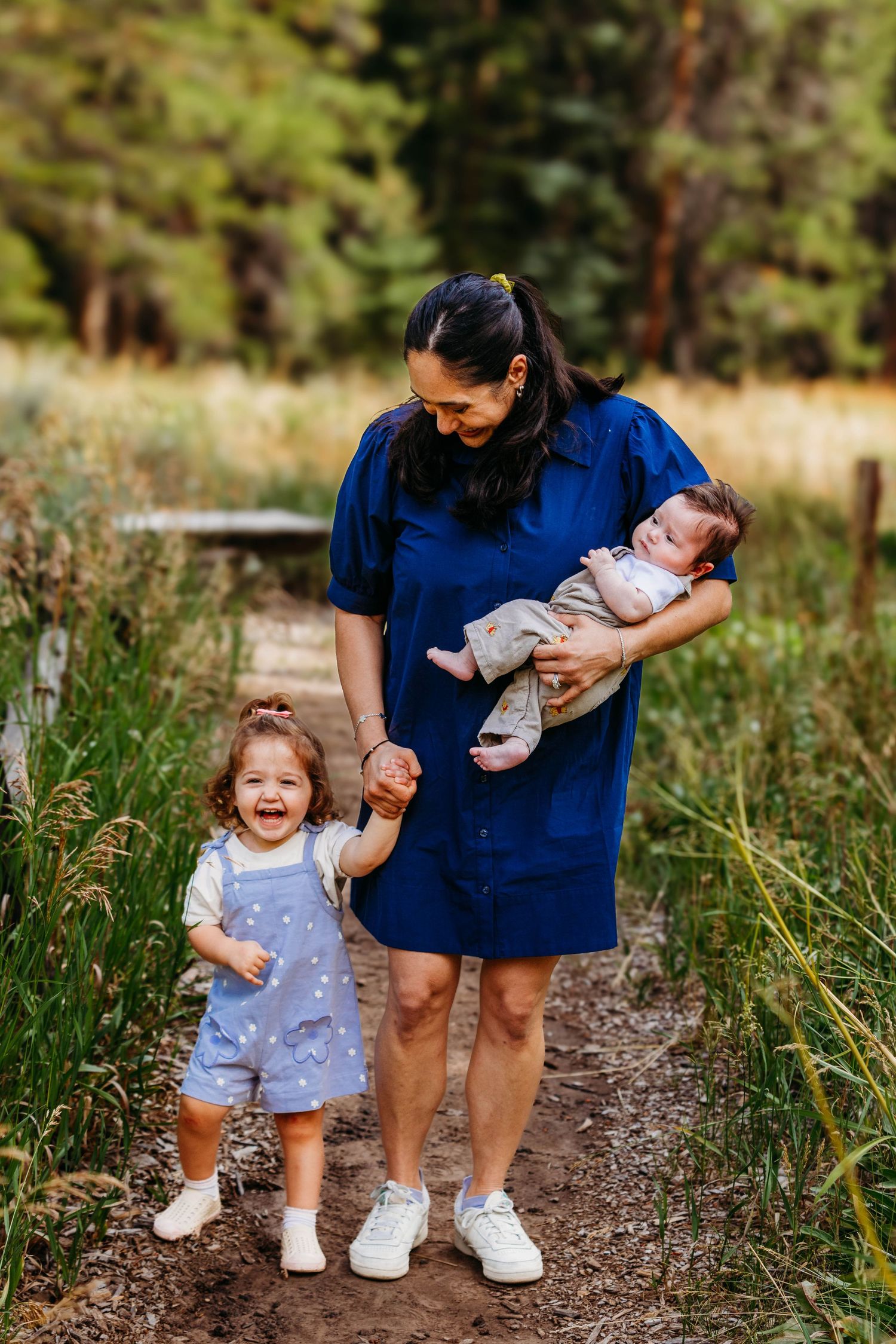 A smiling mother in a blue dress holds a newborn while her toddler daughter laughs on a nature path.