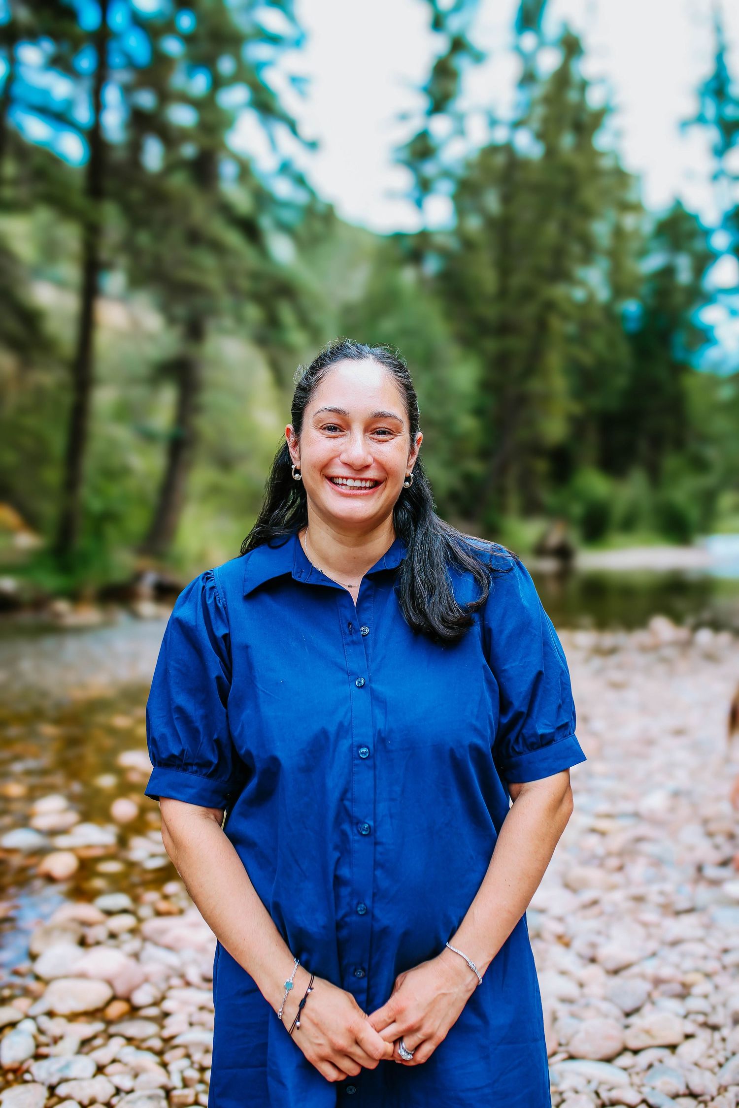 Smiling woman in blue dress standing by a rocky mountain stream surrounded by tall pine trees.