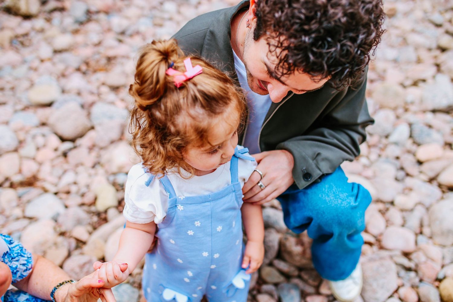 Adult bending down to interact with a toddler girl in blue overalls on a rocky beach.