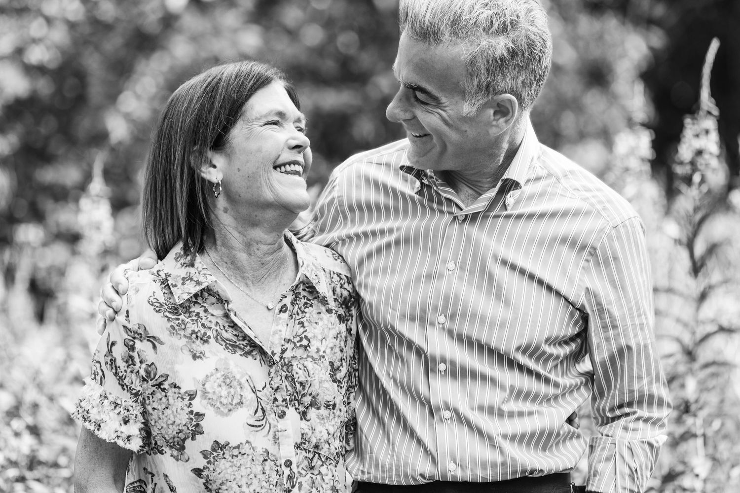 Black and white photo of a smiling middle-aged couple embracing outdoors in a lush garden setting.
