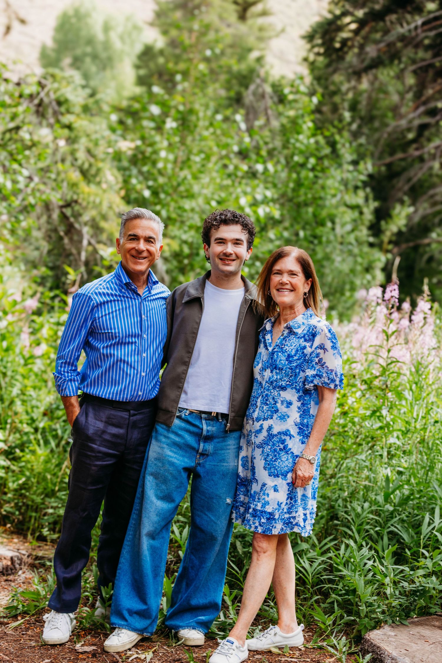 A family of three smiling outdoors surrounded by lush green trees and wildflowers, wearing blue outfits.