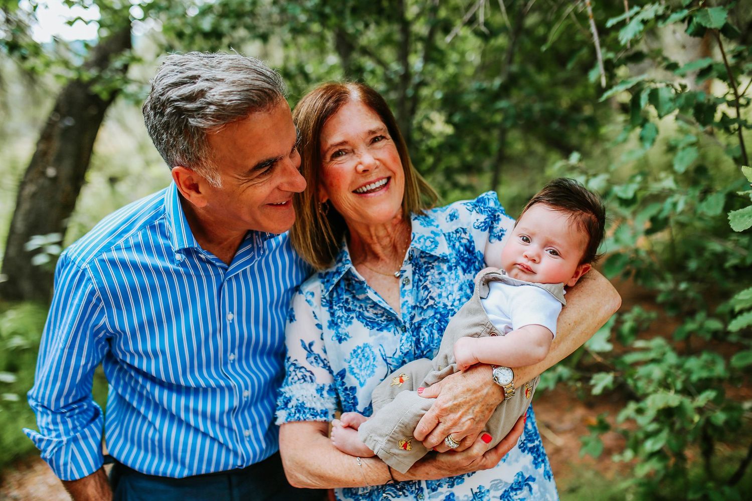 Happy grandparents smiling and holding a baby outdoors in a lush green garden setting.