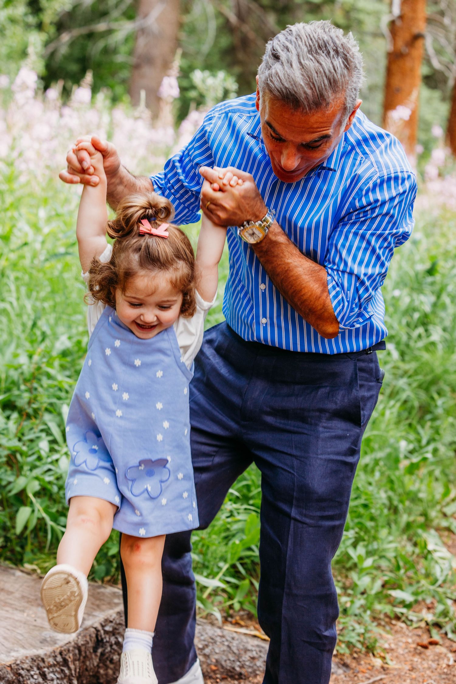 Grandfather in blue striped shirt joyfully helps little girl balance outdoors in lush green garden setting.