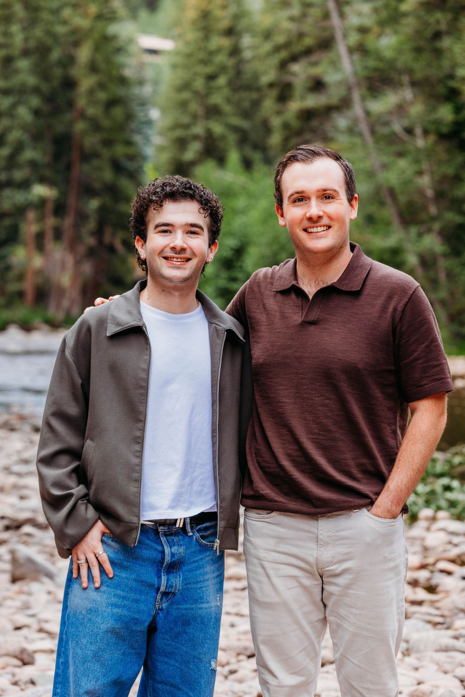 Two smiling young men posing outdoors near a rocky riverbed surrounded by tall green pine trees.