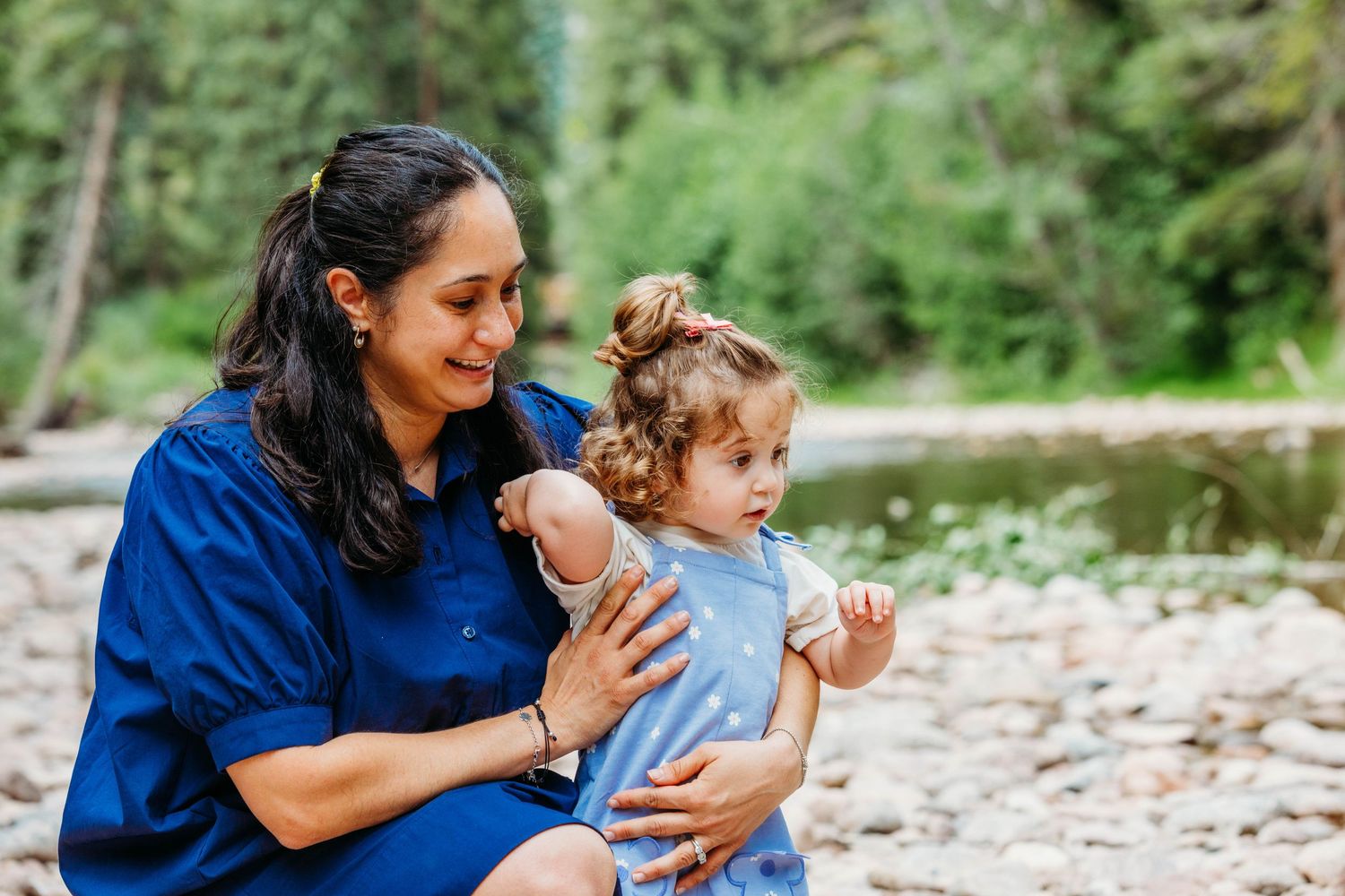 A woman in a blue dress lovingly holds a curly-haired toddler girl outdoors near a rocky riverbed.