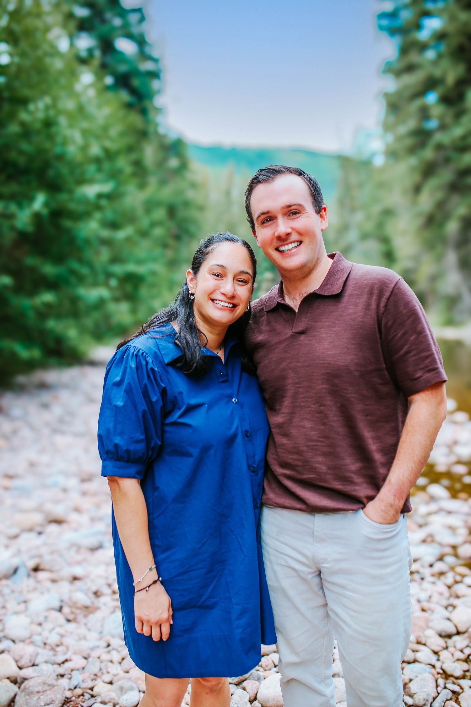 A smiling couple poses together on a rocky riverbank surrounded by tall evergreen trees and mountains.