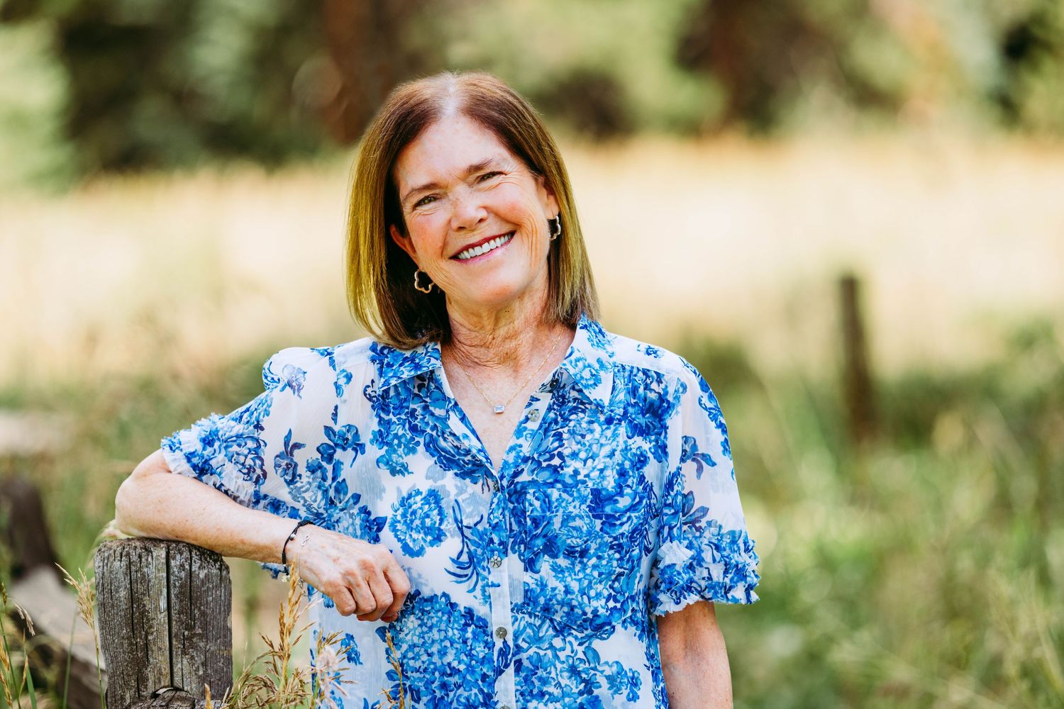 Smiling middle-aged woman in blue floral blouse leaning on wooden post in sunny meadow.