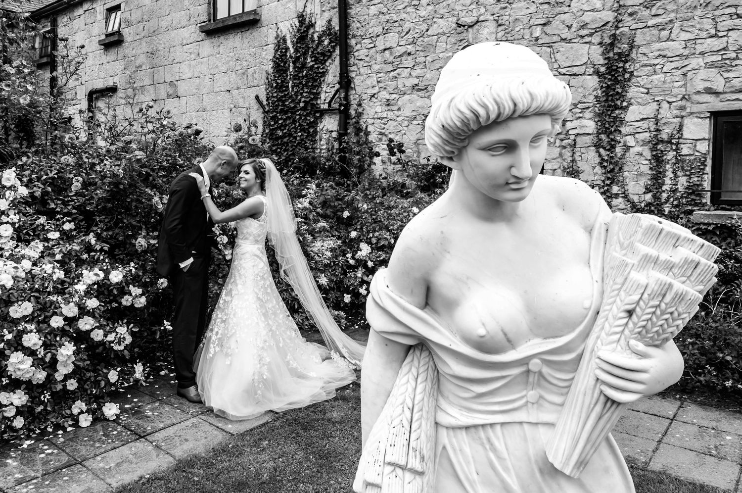 Black and white wedding photo with couple kissing in garden behind classical stone statue at Cabra Castle, Co. Cavan.