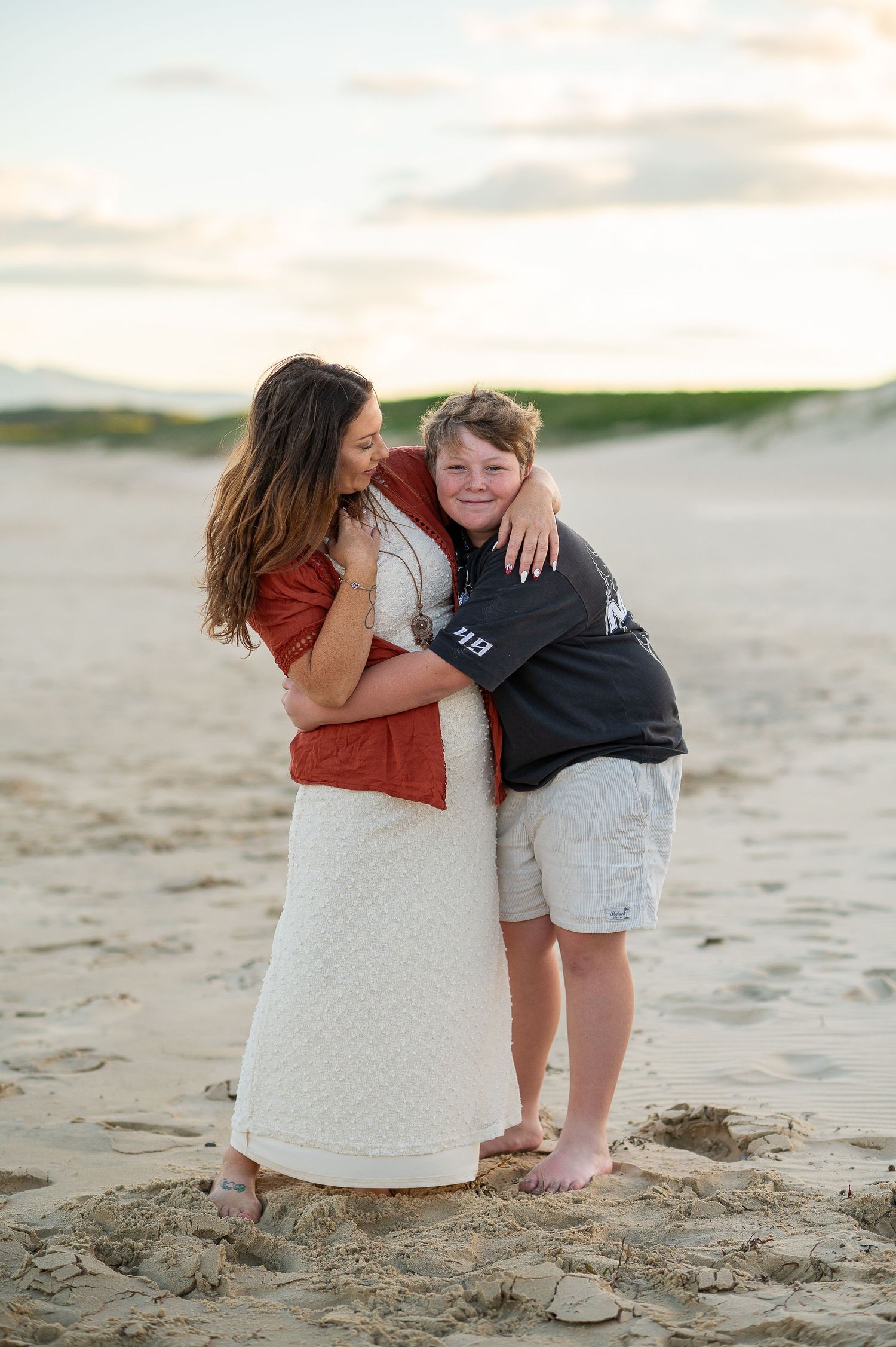A mother and young boy share a warm hug on a sandy beach at sunset, both smiling and barefoot.