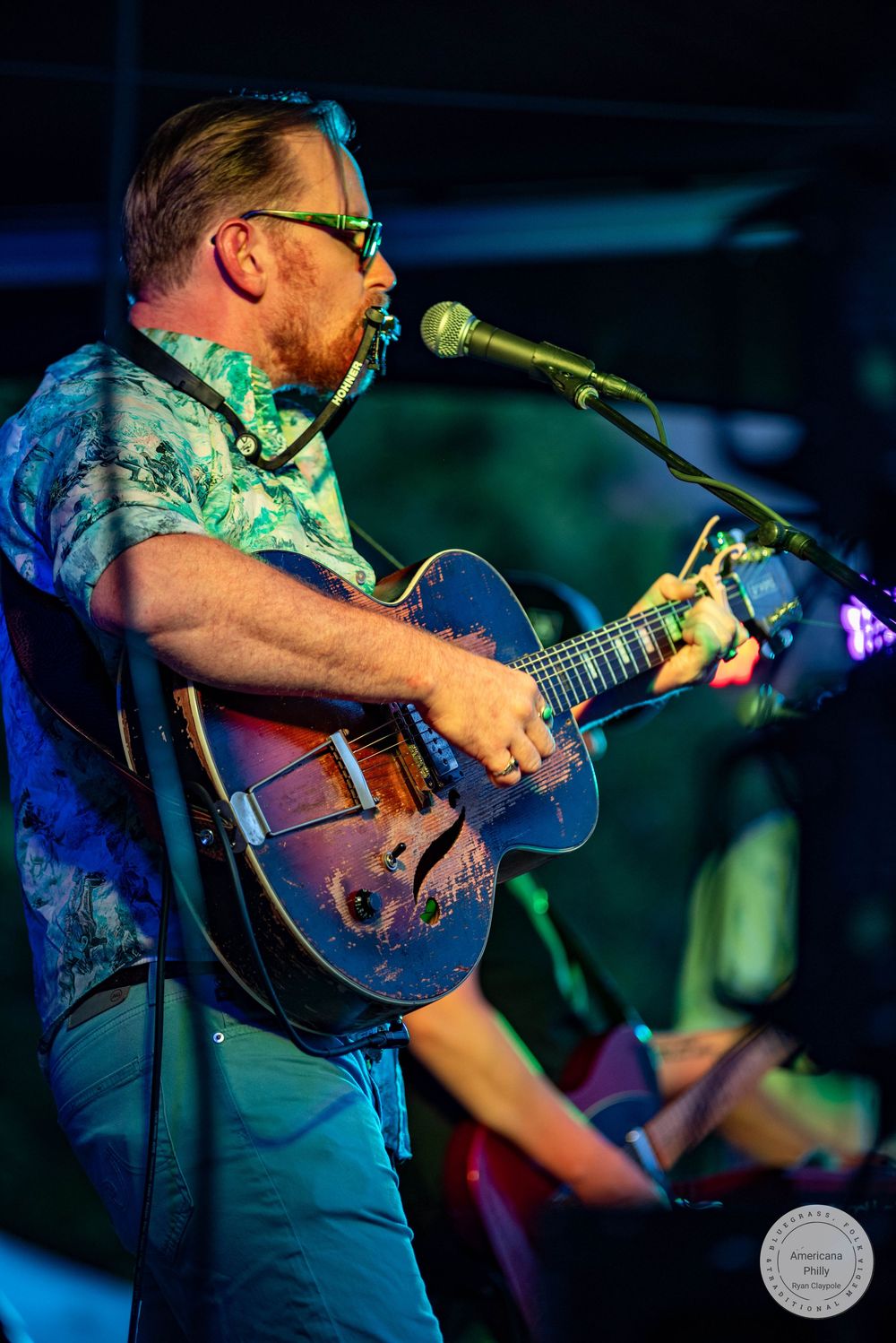 Man with glasses playing archtop guitar and singing into microphone on stage in colorful Hawaiian shirt under blue stage lights.