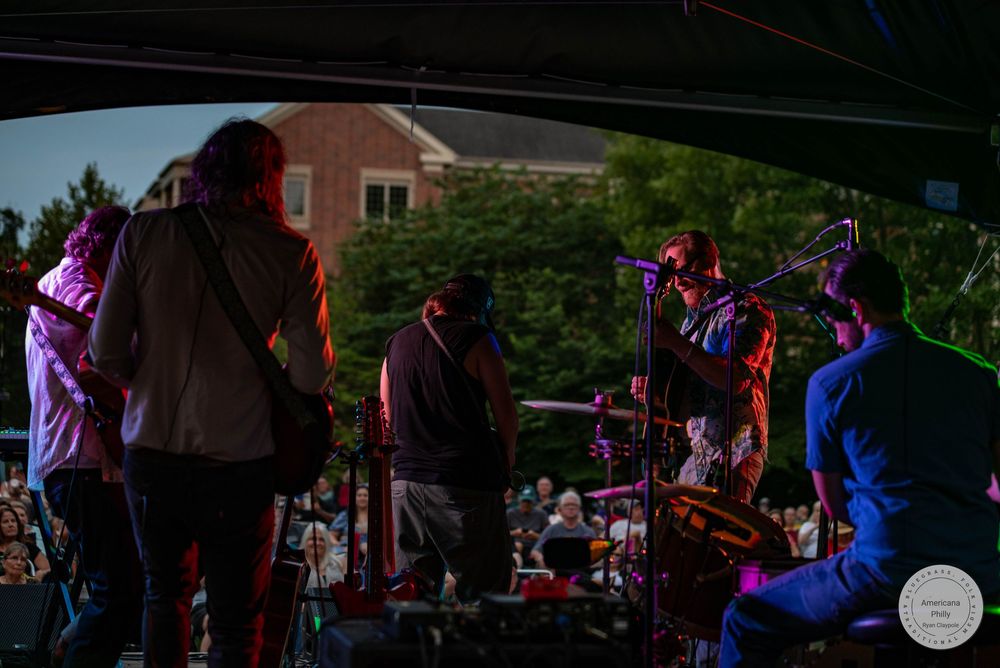 Live band performing on an outdoor stage at dusk with colorful lighting and a crowd gathered in the background.