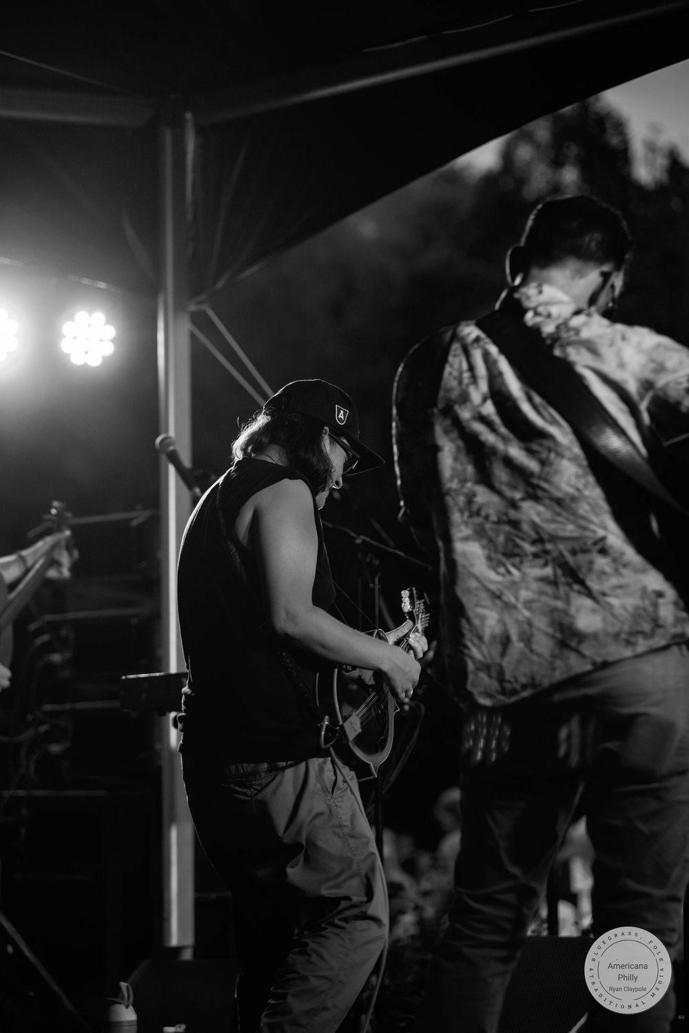 Black and white photo of two musicians performing on stage, one playing guitar under bright stage lights at an outdoor concert.