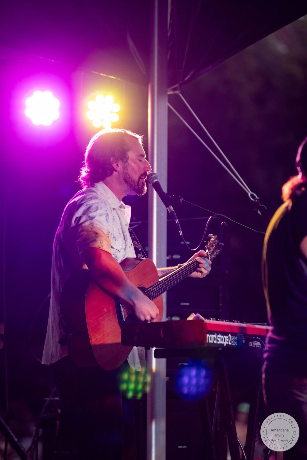 Male guitarist with tattoos performs on stage under colorful purple and orange stage lights at an outdoor concert.