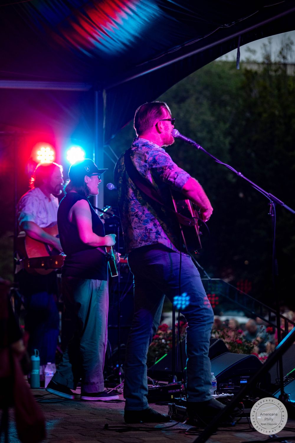 Band performing on outdoor stage under blue and red lights with guitarist at microphone.