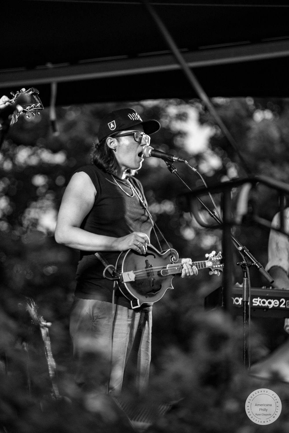 Female musician singing into microphone while playing mandolin on stage at an outdoor concert in black and white photo.