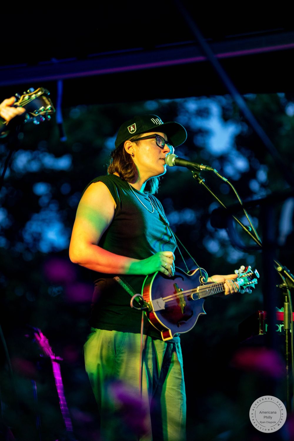 Musician playing mandolin and singing into microphone on outdoor stage at night under colorful lights.