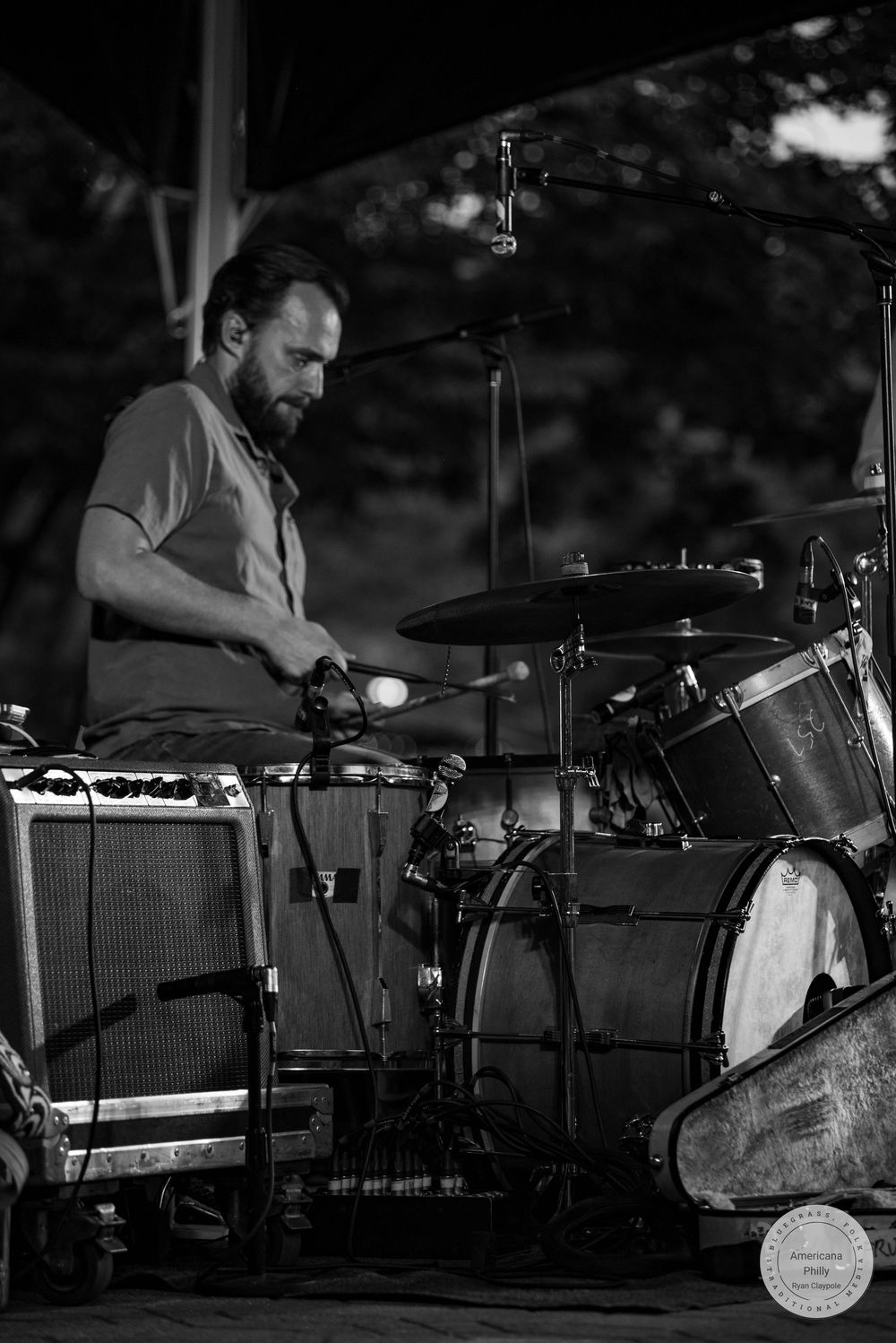 Black and white photo of a bearded male drummer playing a drum kit on an outdoor stage during a live performance.