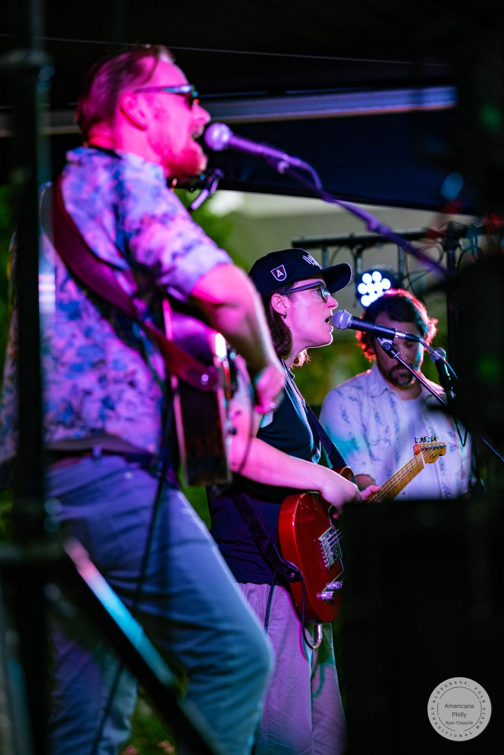 Three musicians perform on stage at night, with guitarist in floral shirt singing into mic under purple stage lights.