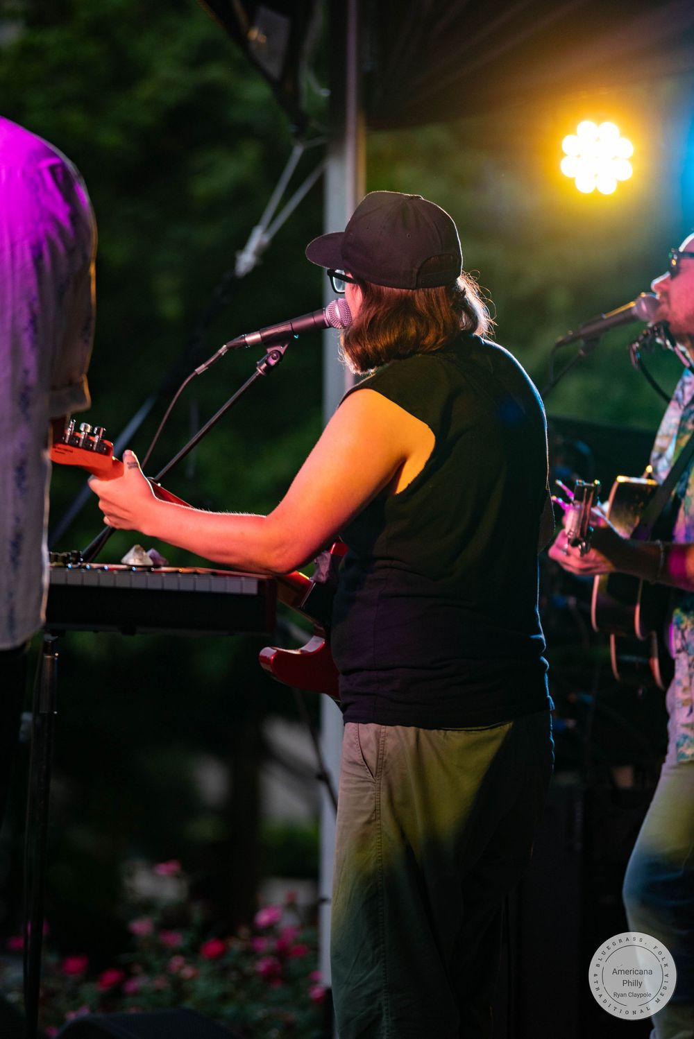 Musician playing keyboard on outdoor stage at night, wearing black cap and top, with stage lighting and guitarist behind.