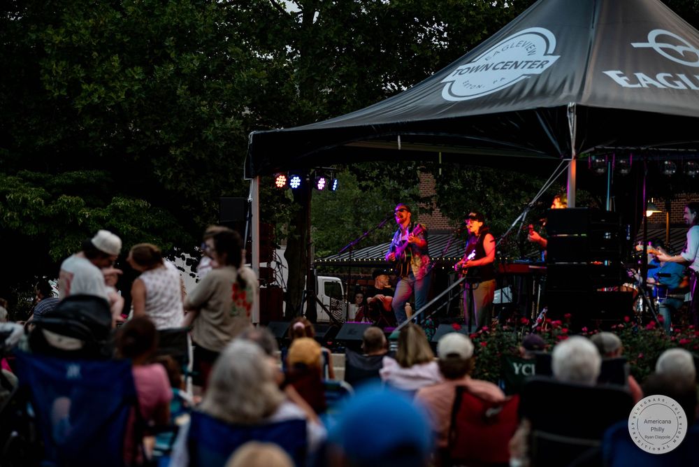 Outdoor evening concert with a band performing under a black canopy tent while a crowd enjoys the live music event.