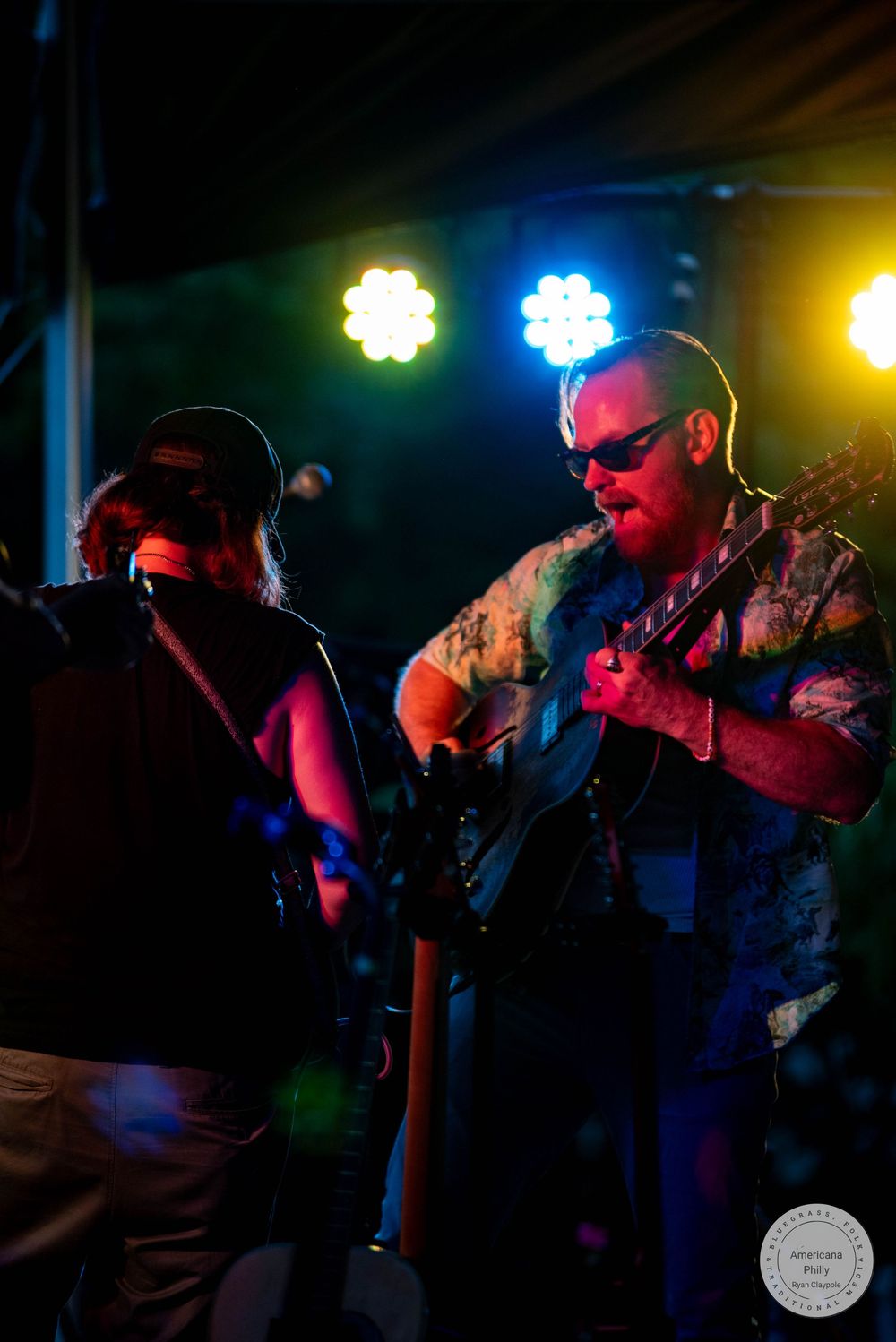 Two musicians performing on stage under colorful blue and red lights, one playing guitar at an outdoor night concert.