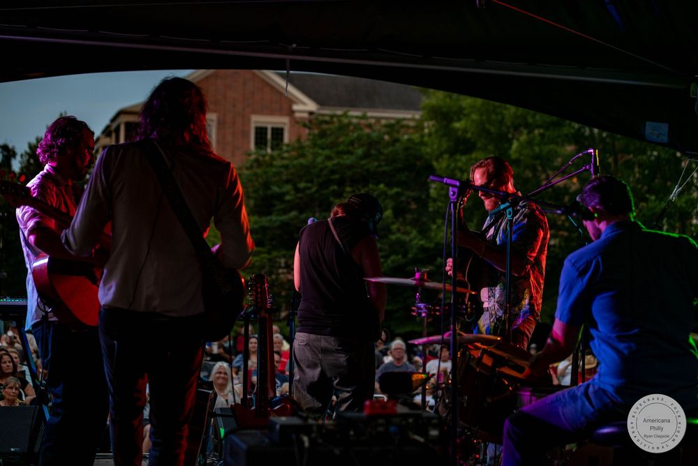 Band performing on an outdoor stage at dusk with audience watching under string lights.