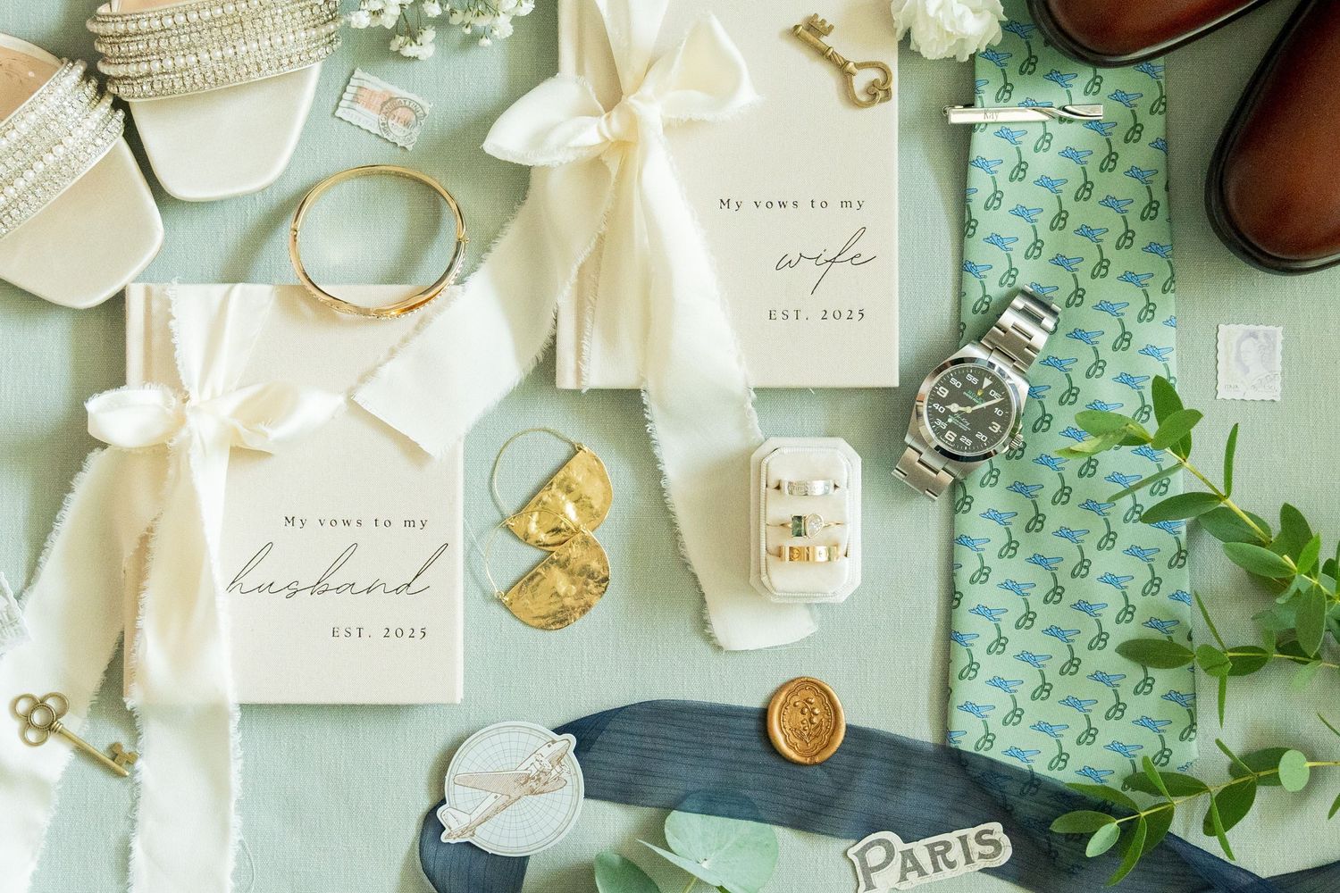 Flat lay of wedding vow books, gold jewelry, a watch, rings, and floral accents on a mint green background.