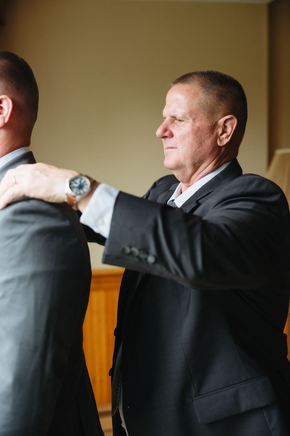 An older man in a dark suit adjusts the jacket of another man during wedding preparations.