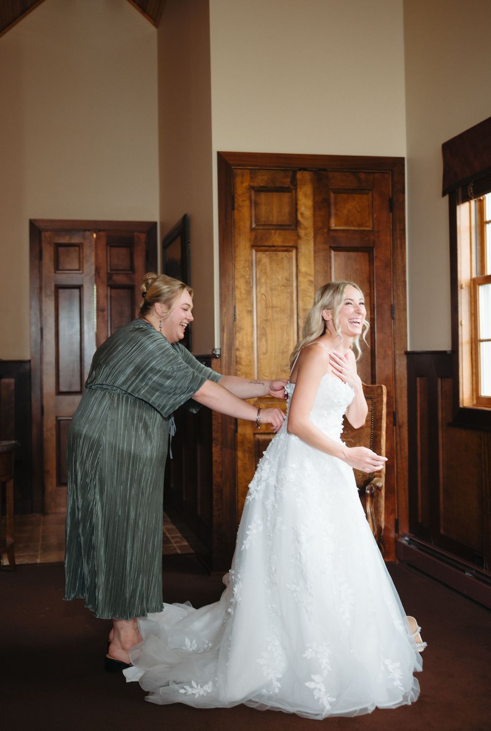 A woman in a green dress helps a smiling bride in a white lace gown get ready inside a rustic wooden venue.
