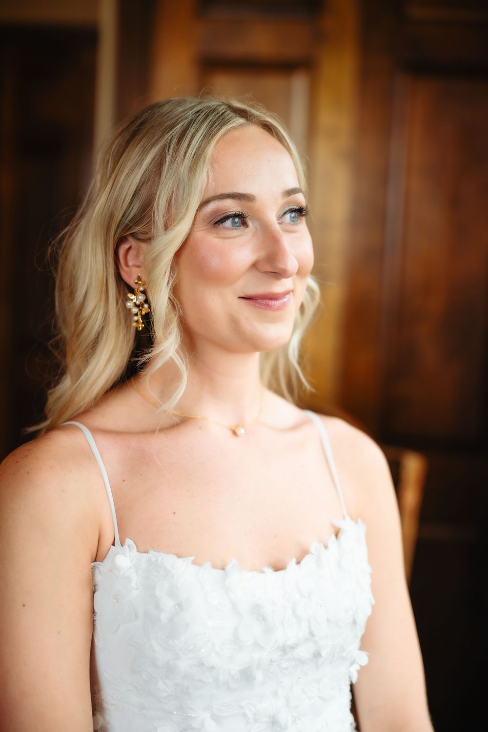 Beautiful blonde bride in white dress wearing gold floral earrings smiling softly on her wedding day.