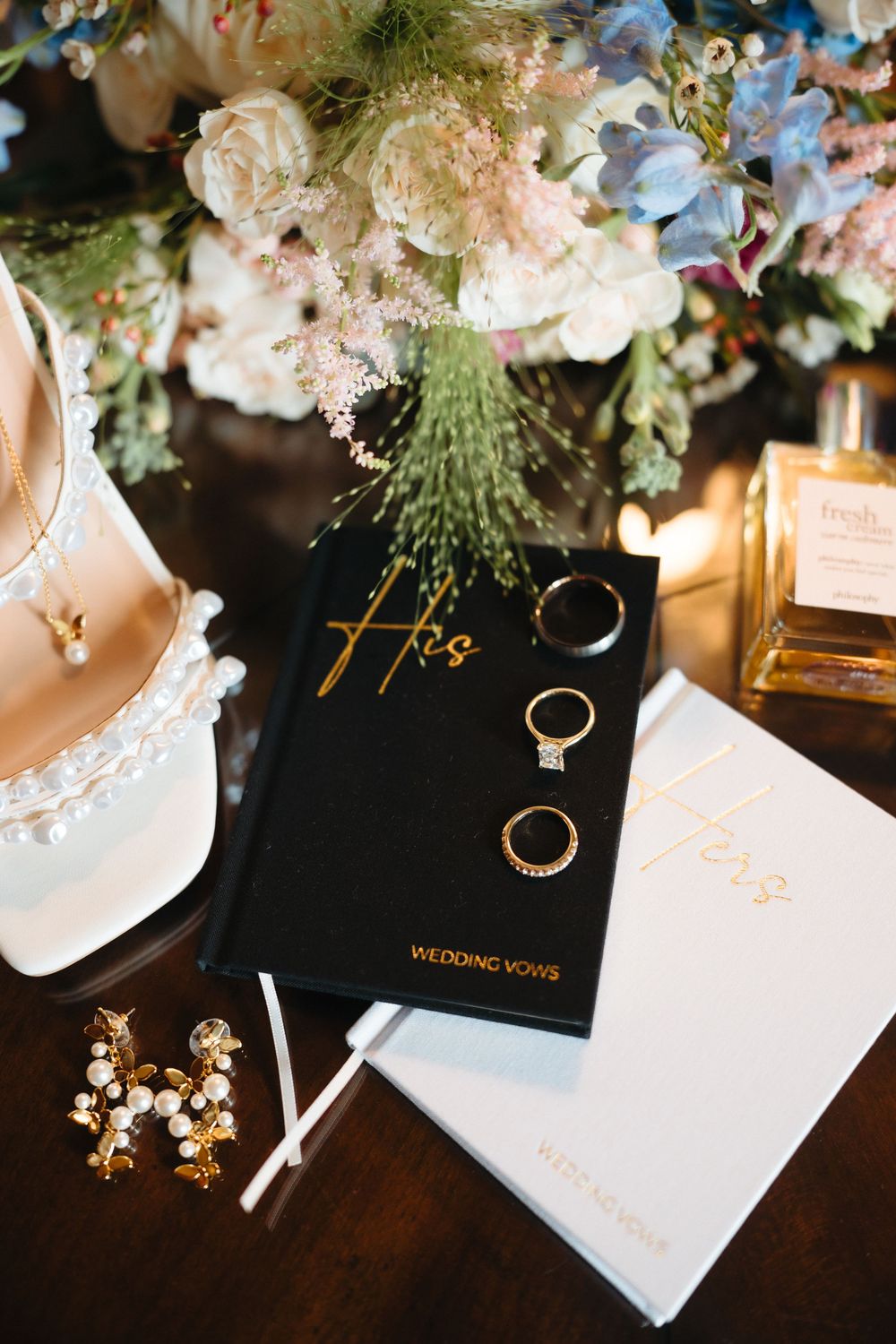 Black wedding vow books with gold rings, pearl earrings, heels, and floral bouquet on dark wood table.